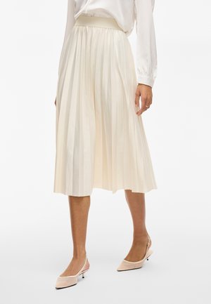 Frau trägt einen elfenbeinfarbenen, plissierten Midirock, kombiniert mit einer weißen Bluse mit langen Ärmeln und beigen Slingback-Schuhen mit niedrigem Absatz.