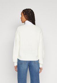 White knitted sweater with a boxy fit and ribbed texture. Ribbed cuffs and hem. Paired with blue wide-leg jeans.