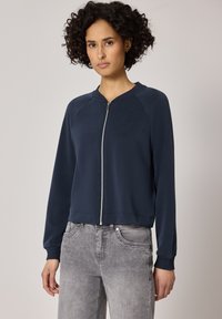 Femme aux cheveux bouclés portant une veste zippée bleu marine et un jean gris clair, debout devant un fond clair uni.