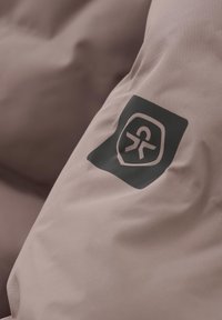 Švelni, tekstūruota medžiaga, neryškiai rožinė, su juodu logotipo pleistru, kuriame pavaizduota prieinamumo simbolis, esantis ant drabužio rankovės.