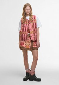 Colete acolchoado rosa com padrão geométrico, usado sobre uma saia com padrão floral. Botins pretos com acentos em rosa. Camisa branca por baixo.