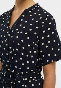 Robe à pois marine avec un col de chemise, des manches courtes, deux poches poitrine et une taille nouée, fabriquée à partir d'un tissu doux avec une texture lisse.