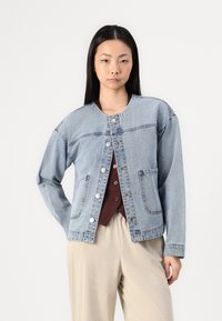 Svetlo modra jeans jakna s okroglo ovratnico, z gumbnim zapiranjem, dvema sprednjima žepoma in sproščenim krojem. Nosimo jo preko vreče rdeče majice in kremno obarvanih hlač.