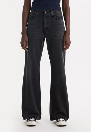 Person iført sorte vidde jeans med høj talje og sorte og hvide sneakers, stående mod en ensfarvet lys baggrund.