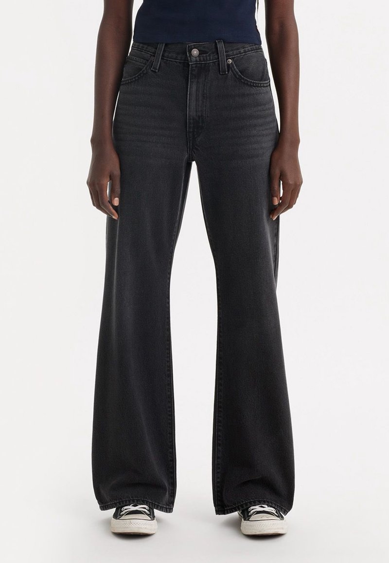 Personne portant un jean noir taille haute à jambes larges et des baskets noires et blanches, debout devant un fond clair uni.