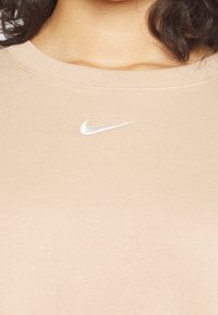 Beżowa koszulka z okrągłym dekoltem na osobie, z białym wyszywanym logo Nike w kształcie łyżwy, umieszczonym centralnie poniżej kołnierzyka.