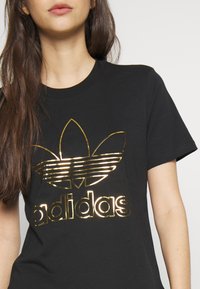 Svart bomulls t-shirt med en glänsande guldig Adidas-logotyp, inklusive trebladsemblem och horisontella ränder, med rund hals och korta ärmar.