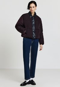 Bordeauxrote Pufferjacke mit schwarzem Kunstleder-Frontpanel und Kragen, in kastiger Form und mit steppenden Details, kombiniert mit dunkelblauen Jeans.
