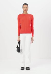 Pull rouge en tricot torsadé avec un col rond, associé à un pantalon blanc évasé et des chaussures noires, tenant un petit sac à main noir.