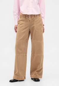 Pantalon large en denim marron clair, avec poches avant, surpiqûres visibles et braguette zippée, associé à une chemise rayée rose.