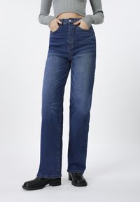 Τζιν μπλε χρώματος με ψηλή μέση και ίσια γραμμή στα πόδια. Διαθέτουν θηλιές για ζώνη, μπροστινές τσέπες και διακριτική φθορά κοντά στα γόνατα.