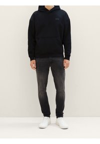 Mikina s kapucí v tmavě námořnické barvě s přední kapsou, spárovaná se sepranými černými džínami a bílými teniskami. Hladká látka, uvolněný střih.