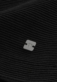 Tessuto nero a costine con dettaglio logo quadrato metallico. La texture è morbida ed elastica, con sottili linee orizzontali.