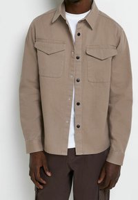 Beżowa koszula na guziki z wytrzymałej tkaniny, z dwoma kieszeniami na piersi, czarnymi guzikami i klasycznym kołnierzykiem. Nosi się ją na białej koszulce.