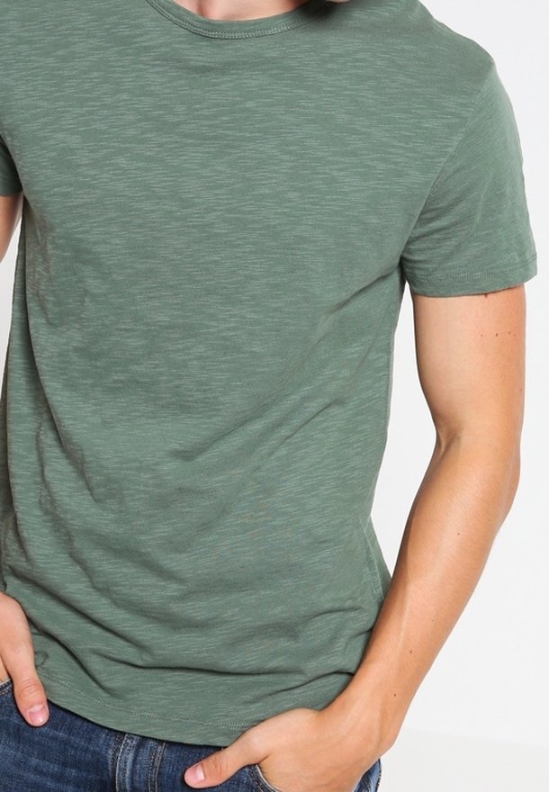 Homme portant un t-shirt vert à manches courtes ajusté et un jean bleu, mains partiellement dans les poches, sur un fond blanc uni.