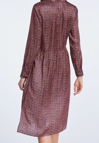 Femme portant une robe à manches longues, longueur genou, avec un petit motif floral rose et noir, vue de dos sur un fond uni.