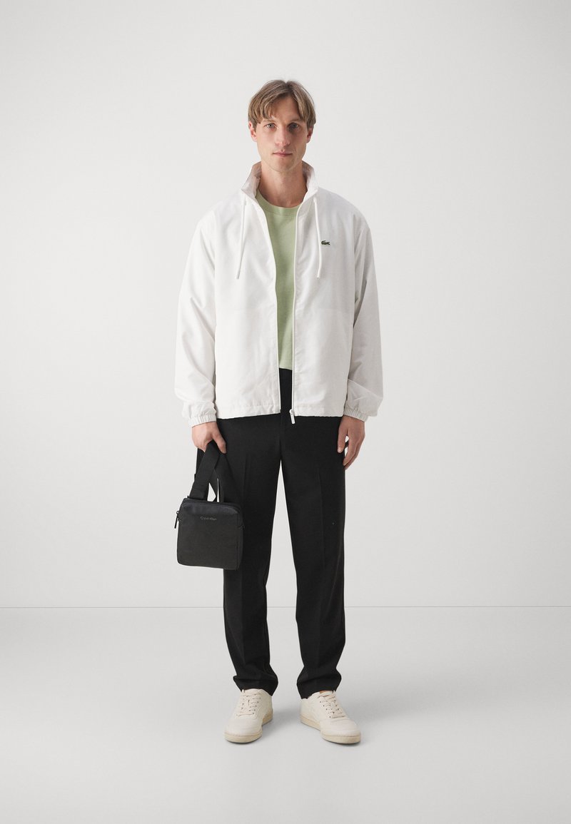 Weiß leichte Jacke mit hohem Kragen, grünes T-Shirt, schwarze Hose und cremefarbene Sneakers; hält eine kleine schwarze Tasche mit einem Riemen.