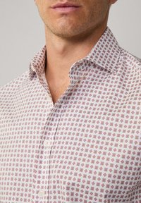 Witte button-up blouse met een klein geometrisch rood patroon. Zachte textuur met een klassieke kraag en twee zichtbare knopen.