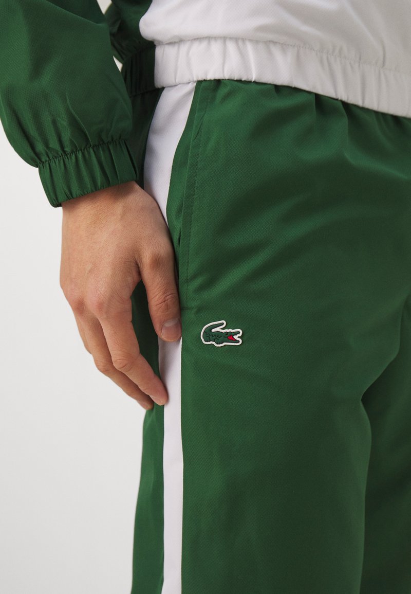 Pantalon de sport vert en tissu léger avec des poignets élastiques et des bandes latérales. Présente un logo de crocodile sur la cuisse.