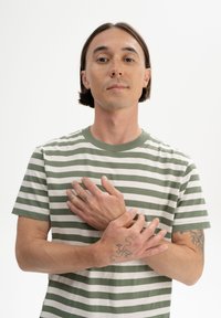 Grün-weißer gestreifter T-Shirt mit Rundhalsausschnitt. Das Modell hat kurze Haare und zeigt mehrere sichtbare Tattoos an Händen und Armen.