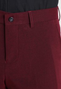 Burgundy színű nadrág, texturált anyagból készült, zipzárral, gombos záródással, övlyukakkal és egy oldalsó zsebbel.