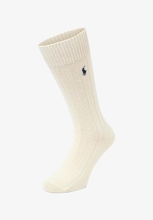 Chaussettes côtelées montant jusqu'au genou de couleur crème avec une texture douce. Ornées d'un logo polo bleu marine sur le côté. Lignes verticales régulièrement espacées sur toute la longueur.