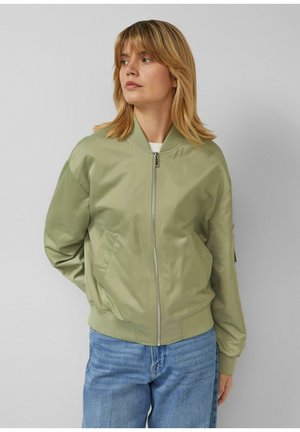 Blonde Frau trägt eine hellgrüne Bomberjacke mit Frontreißverschluss und blaue Jeans, blickt nach links vor einem einfarbigen Hintergrund.