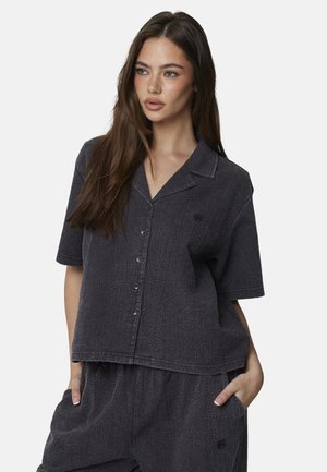 Vrouw met lang bruin haar, gekleed in een donkergrijs, gestructureerd, kortermig overhemd met knopen en bijpassende broek, met handen in de zakken.