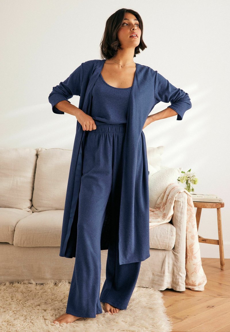 Tamsiai mėlynas poilsio drabužių komplektas, kuriame yra ilgas kardiganas, maikutė ir plačios kelnės, pagaminti iš minkštos medžiagos. Sėdi ant šviesiai smėlio spalvos sofų su šviesia dekoracija.