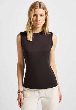 Blonde vrouw in een mouwloze zwarte top en witte broek, staande tegen een effen witte achtergrond, met een zilveren armband om haar linkerpols.