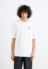 T-shirt en coton blanc à manches courtes, avec un logo noir de Jordan sur la poitrine gauche. Associé à un pantalon texturé foncé avec des accents rouges.