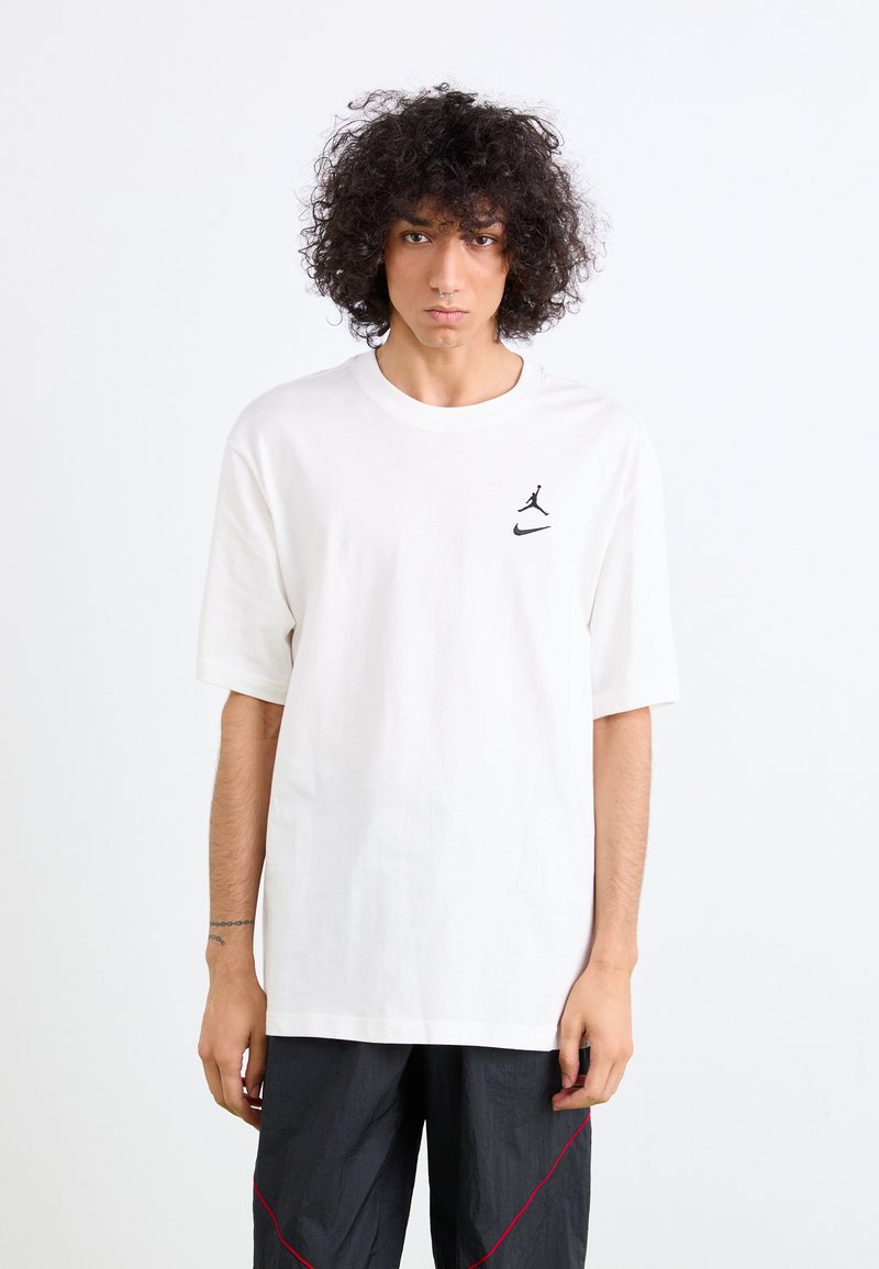T-shirt de algodão branca com mangas curtas, apresentando um logotipo preto da Jordan no lado esquerdo do peito. Combinada com calças escuras, texturizadas, com detalhes em vermelho.