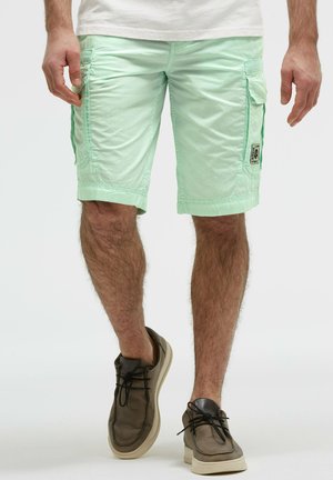 Mann trägt hellgrüne Cargo-Shorts und braune Freizeitschuhe, steht vor einem schlichten Hintergrund.