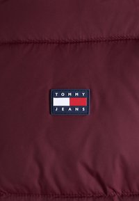Burgunderfarbene Pufferjacke mit strukturierter Oberfläche, ausgestattet mit einem rechteckigen, marineblauen Etikett, auf dem "TOMMY JEANS" in weißen und roten Blockbuchstaben steht.