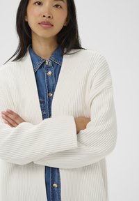 Žena s dlhými tmavými vlasmi v bielej rebrovanej kardigánovej bunde, oblečenej na modrej košeli s gombíkmi, s prekríženými rukami na svetlom pozadí.