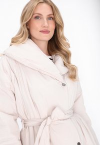 Palton matlasat roz deschis cu guler supradimensionat, talie evidențiată cu curea și accente cu nasturi. Textură netedă și croială lejeră, potrivit pentru a fi purtat pe sub alte haine.