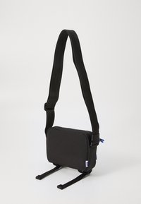 Crni crossbody ranac pravokutnog oblika, sa zatvaračem na zatvarač, podesivim remenom i plavim naglaskom na zatvaraču. Tkanina je glatka.