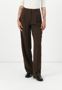 Pantaloni a pieghe marroni con vita alta, gambe larghe e una superficie liscia, abbinati a scarpe nere.