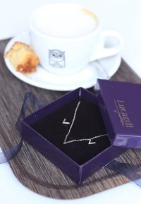 Zilveren ketting met V-vormige hangers in een paarse juwelendoos, liggend op een houten dienblad naast een kopje koffie en een stuk taart.