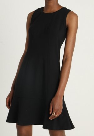 Sleeveless black knee-length dress with a round neckline, worn by a person with medium-dark skin tone, showing right arm and hand with rings.