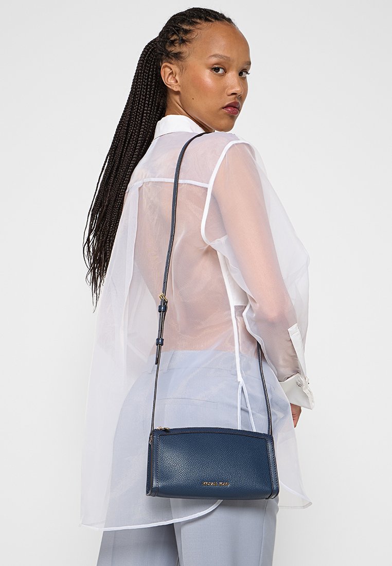 Femme avec de longues tresses portant une blouse blanche transparente et un pantalon gris clair, portant un sac bandoulière bleu marine sur son épaule.