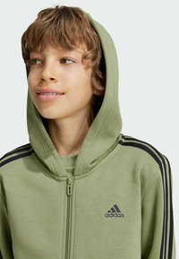 Sudadera verde con cremallera, hecha de material suave y con capucha. Presenta detalles de rayas negras en las mangas y el logo de Adidas en el pecho.