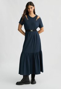 Abito lungo blu navy con maniche corte, dettagli sulle spalle cut-out, orlo a balze e una cintura nera con fibbia argentata. Indossato con stivali neri.