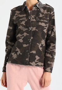 Kvinna bär en mörkgrön och brun camouflagemössa med knäppning samt ljusrosa byxor.