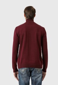 Maronenfarbener Rollkragenpullover mit geripptem Kragen und Bündchen, aus einem weichen Strickmaterial hergestellt. Getragen mit blauen Jeans mit auffälligen Gesäßtaschen.