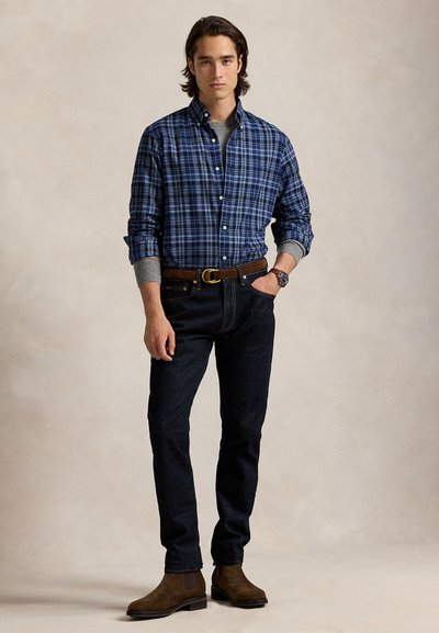 Fiatal férfi, kék kockás ingben, szürke pulóver fölött, sötét farmerben, barna övvel, barna csizmában és karórával, egyik kezét a zsebében tartva áll.