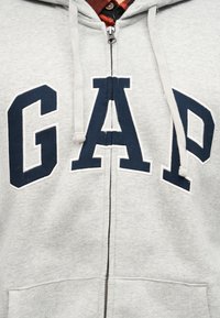 Sweat à capuche gris clair avec fermeture éclair, lettres "GAP" bleu marine et cordons blancs, superposé à une chemise à carreaux rouge et noir.