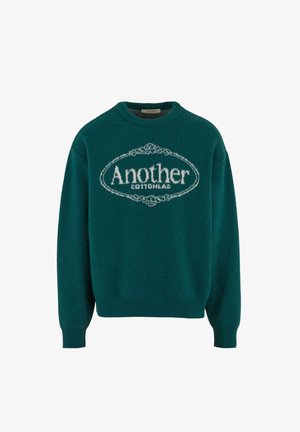 Zeleno pleteno pulover s okroglo ovratnikom, ki ima bel tekst s napisom "Another COTTONLAB" v teksturiranem dizajnu. Ribušeni manšeti in rob.