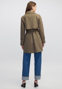 Trench coat caqui con un diseño recortado, cintura ceñida con cinturón, puños abotonados y pliegues en la parte posterior, combinado con jeans azules con dobladillo rodante.