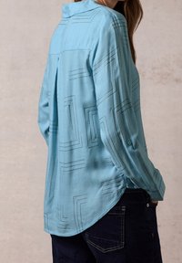 Camicia azzurra a maniche lunghe realizzata in tessuto morbido, con un motivo geometrico in blu scuro, polsino con bottone e dettaglio della piega posteriore.